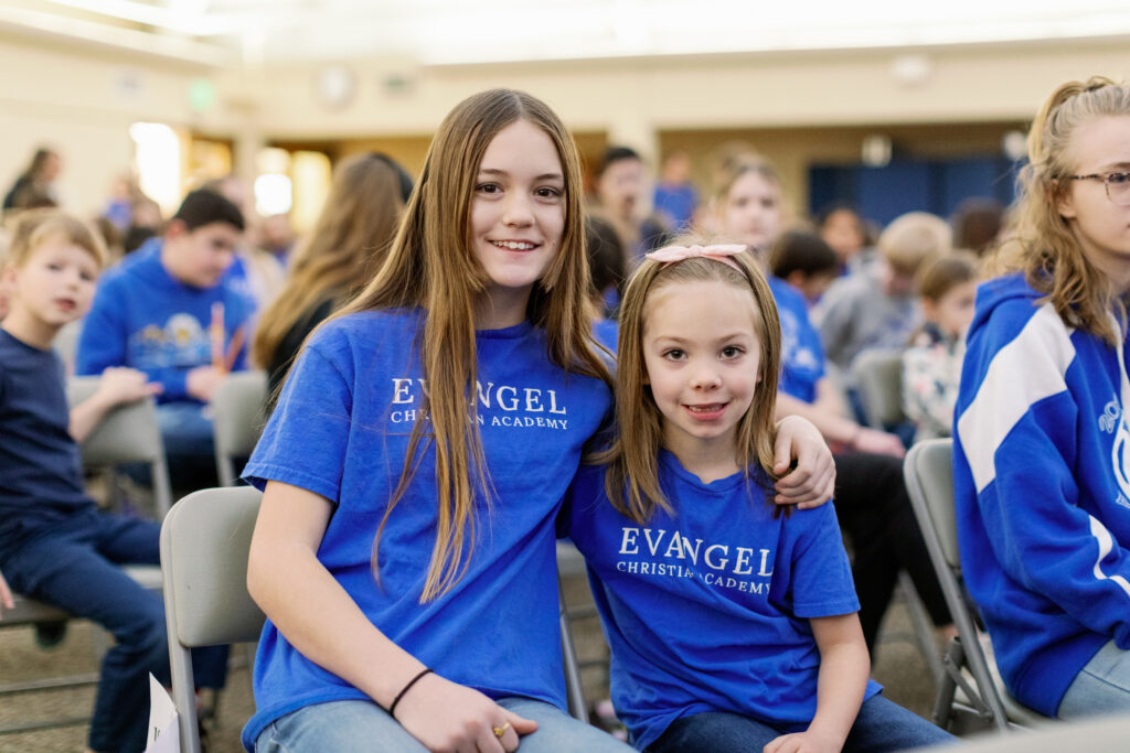 A high school girl and an elementary girl sitting together at a school chapel service. 