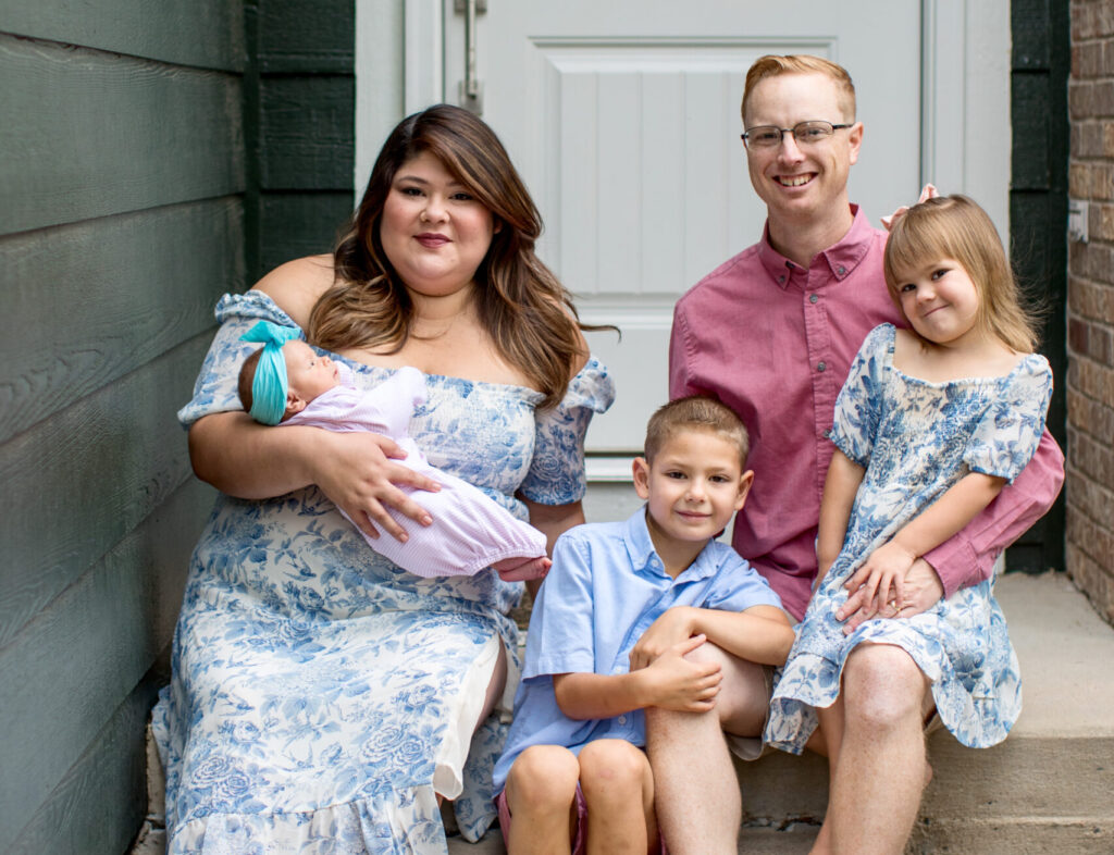 A mom, dad, teenage son, elementary daughter, and preschool son stand together outside of a brick house. 